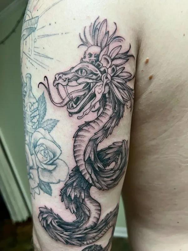 Black-and-grey tattoo of a feathered serpent with a skull headdress coiling down an upper arm