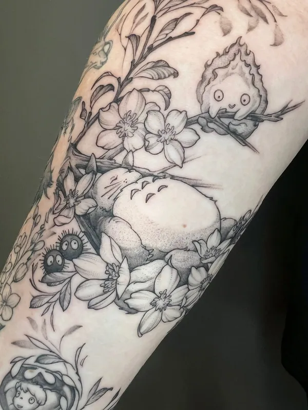 Black-and-grey Studio Ghibli tattoo of a sleeping Totoro, Calcifer and soot sprites among cherry blossoms on a forearm