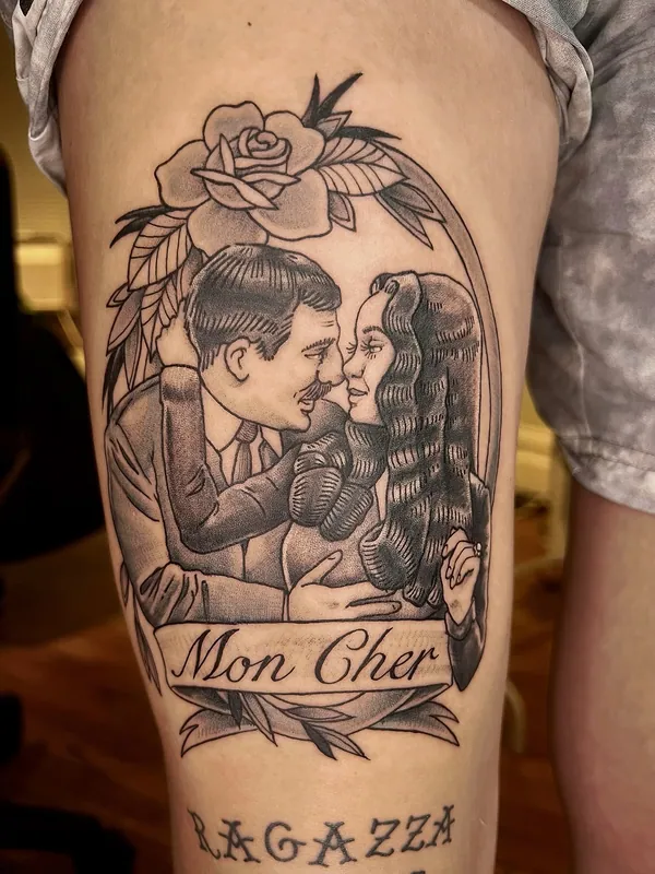 Black-and-grey tattoo of Gomez and Morticia Addams nose to nose under a rose, with a banner reading 'Mon Cher', on a thigh