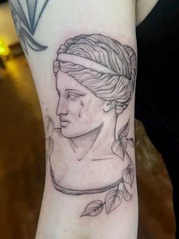 Fine-line black-and-grey tattoo of a classical marble bust with a single tear, ringed by a leafy vine, on an inner upper arm