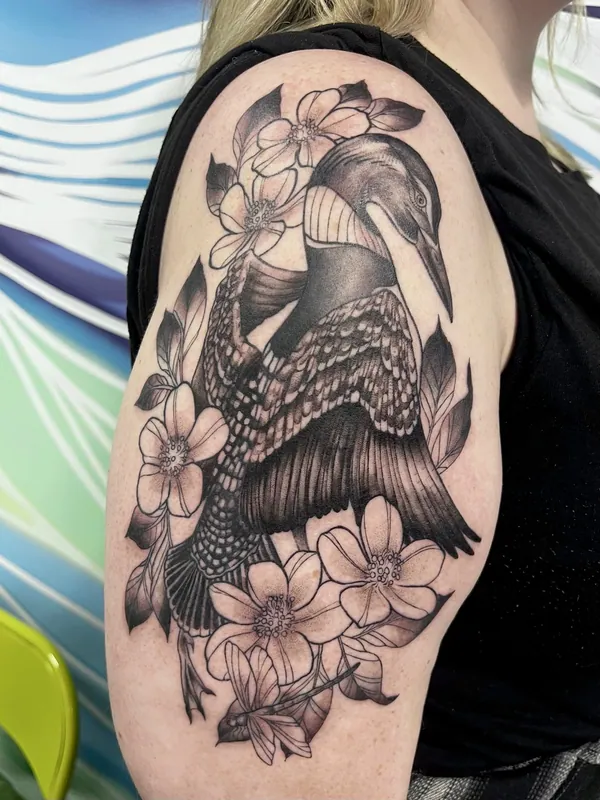 Black-and-grey tattoo of a common loon with checkered plumage among dogwood blossoms on an upper arm