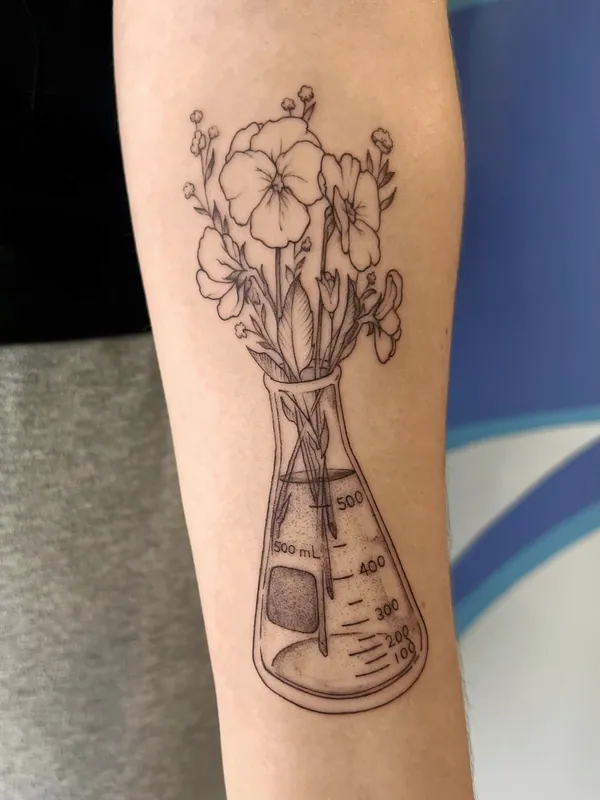Fine-line black-and-grey tattoo of pansies and wildflowers arranged in a 500 mL Erlenmeyer flask on a forearm