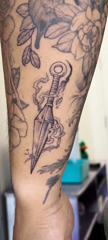 Black-and-grey tattoo of a kunai knife wreathed in smoke on a forearm, between older peony and animal tattoos