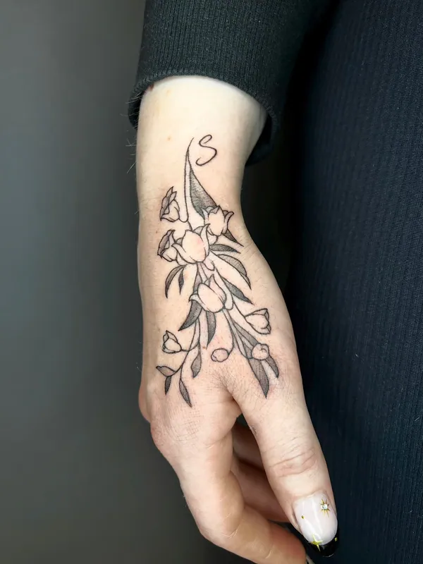 Fine-line black-and-grey tulip tattoo with leaves and a small initial on the back of a hand and wrist