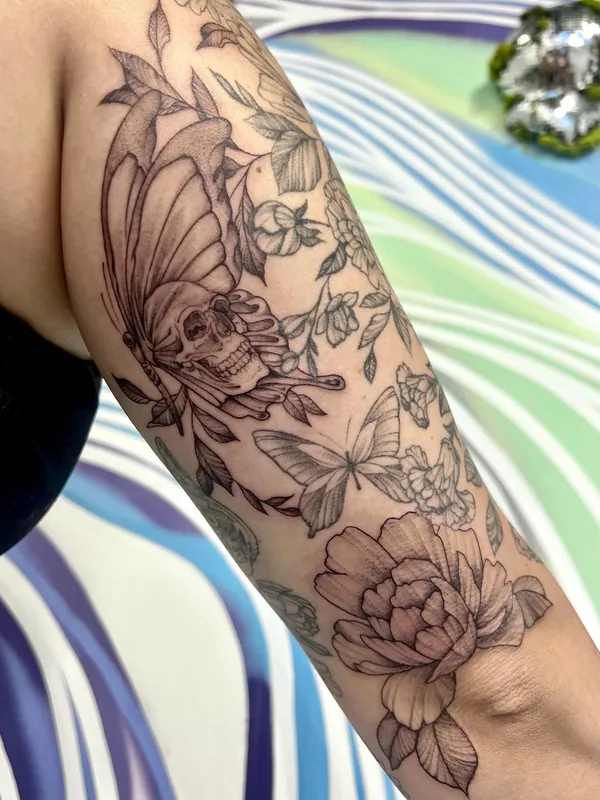 Black-and-grey sleeve tattoo of a skull-headed butterfly, a second butterfly, blossoms and a peony running down a forearm