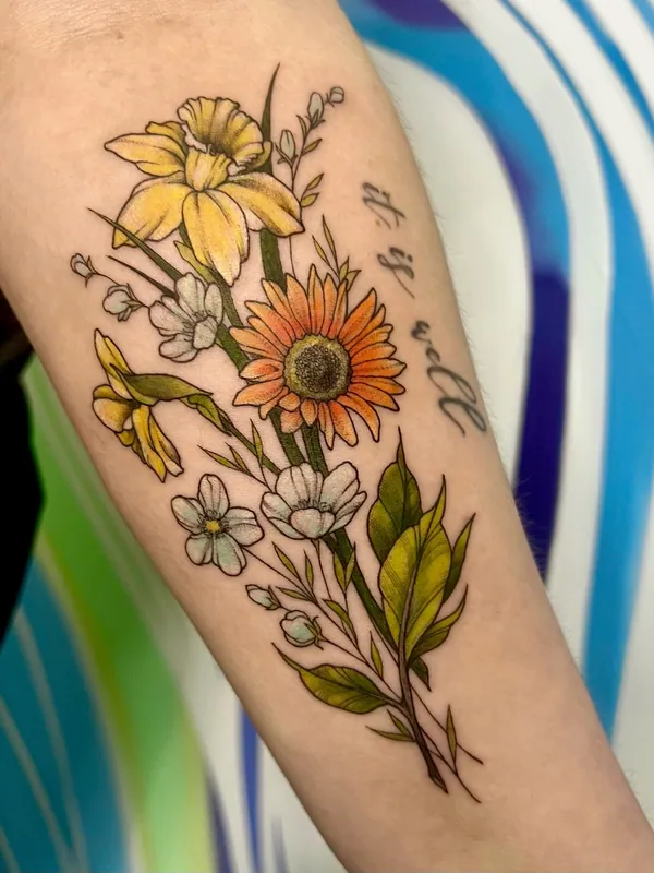 Color tattoo of daffodils, an orange gerbera and white wildflowers with the script 'it is well' on a forearm