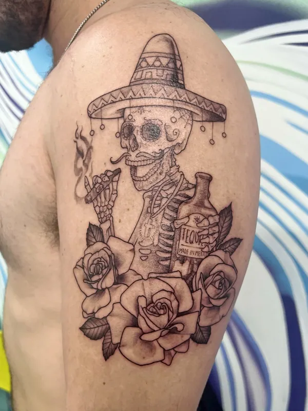 Black-and-grey tattoo of a skeleton in a sombrero smoking a cigar beside a tequila bottle and roses on an upper arm