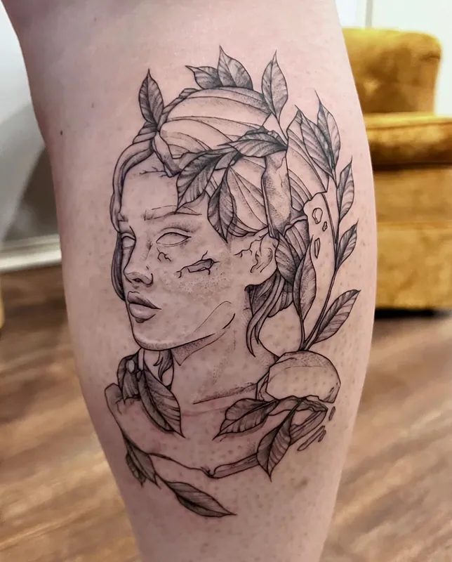 Fine-line black-and-grey tattoo of a cracked statue-like face with leaves growing through it on a calf