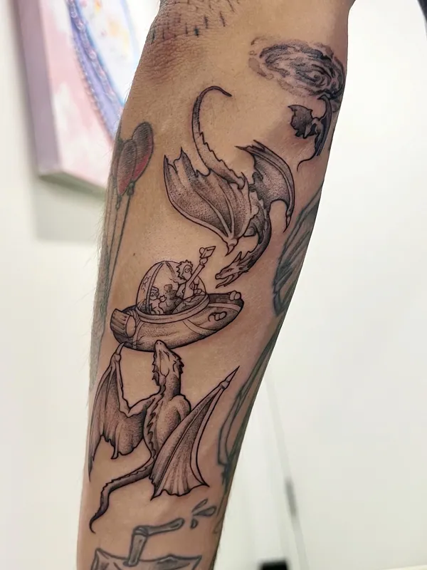 Black-and-grey fine-line tattoo of two dragons flying around a small spaceship with a figure aboard on a forearm