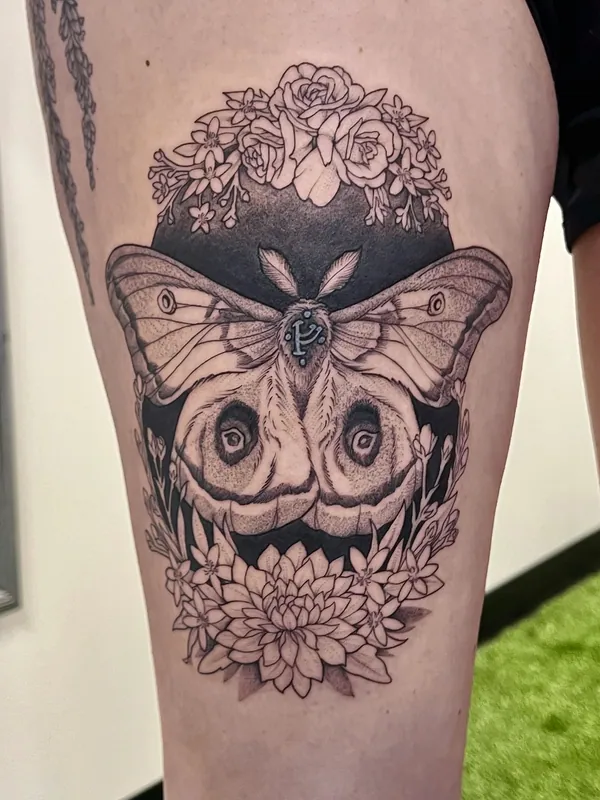 Black-and-grey tattoo of an emperor moth with eyespot wings on a black ground framed by roses and a chrysanthemum on a thigh