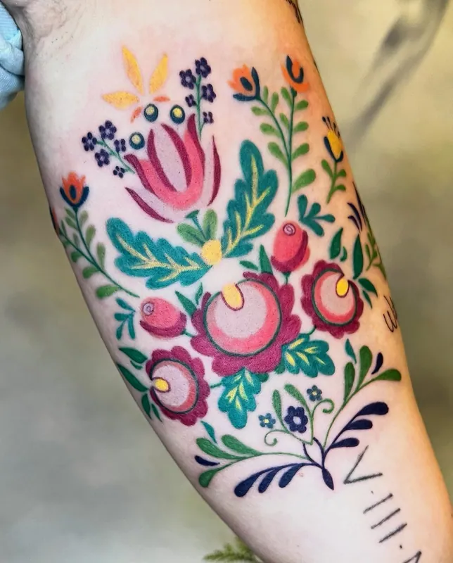 Bold color folk-art floral tattoo in pinks, greens and yellows in the style of Polish papercuts on a forearm