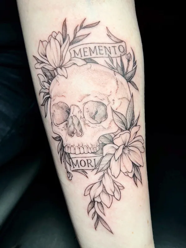 Fine-line black-and-grey skull tattoo with lilies and banners reading 'Memento Mori' on a forearm