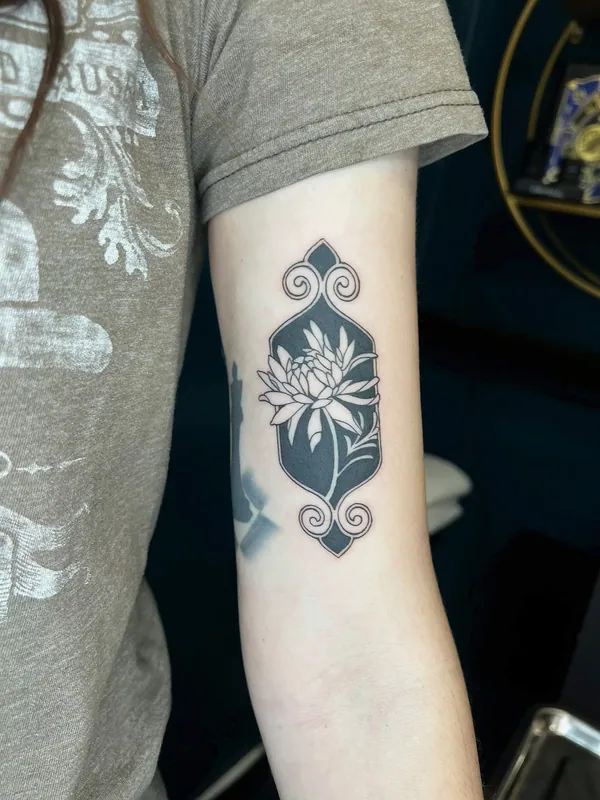 Blackwork tattoo of a chrysanthemum in negative space inside an ornamental scrolled frame on an upper arm