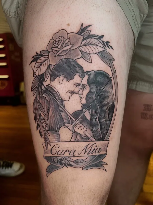Black-and-grey tattoo of Gomez and Morticia Addams embracing under a rose with a banner reading 'Cara Mia' on a thigh