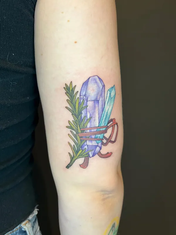 Color tattoo of two crystals bound to a sprig of rosemary with red ribbon on the back of an upper arm
