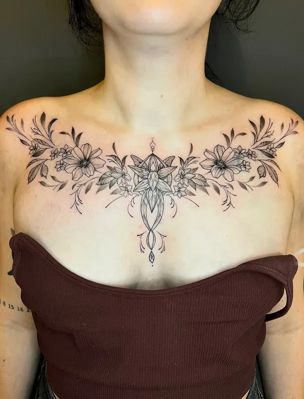 Symmetrical fine-line black-and-grey chest tattoo of wildflowers and leaves spreading from an ornamental sternum pendant