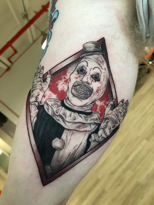 Tattoo of Art the Clown from Terrifier grinning inside a diamond frame with red color splatter on an inner upper arm