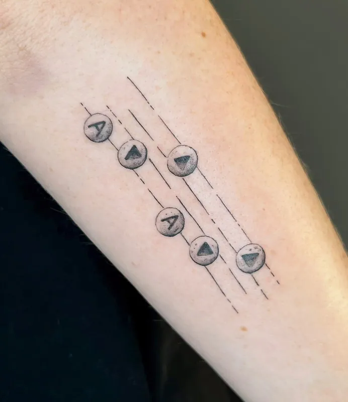 Minimal dotwork tattoo of six round buttons marked A and play symbols strung along thin lines on a forearm
