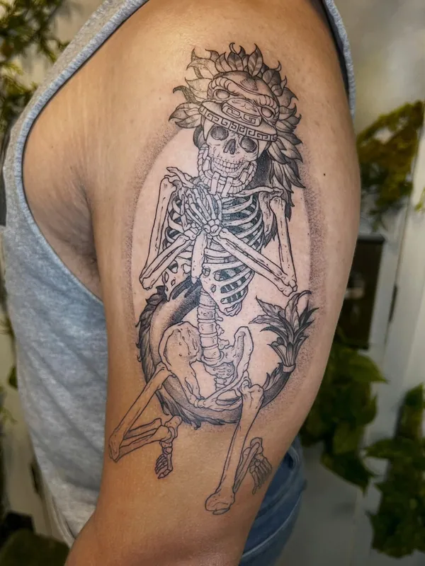Black-and-grey fine-line tattoo of a seated skeleton wearing a feathered Aztec headdress on an upper arm