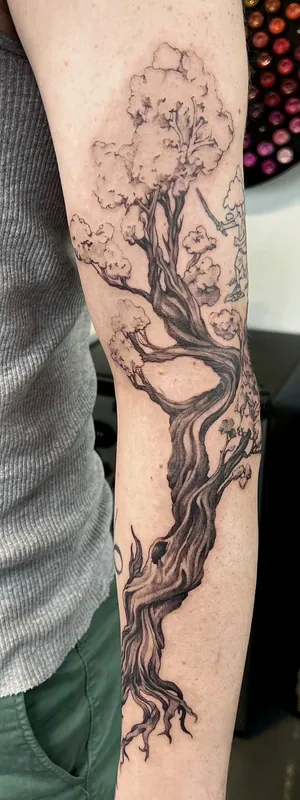 Black-and-grey tattoo of a gnarled blossoming tree with exposed roots running the full length of a forearm
