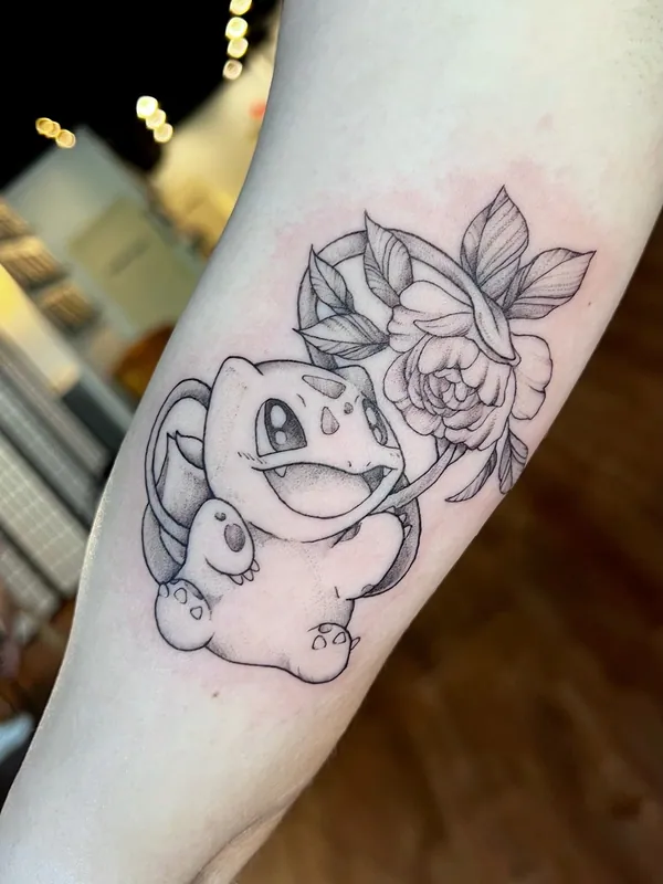 Fine-line black-and-grey tattoo of Bulbasaur from Pokemon beside a blooming rose on an inner forearm