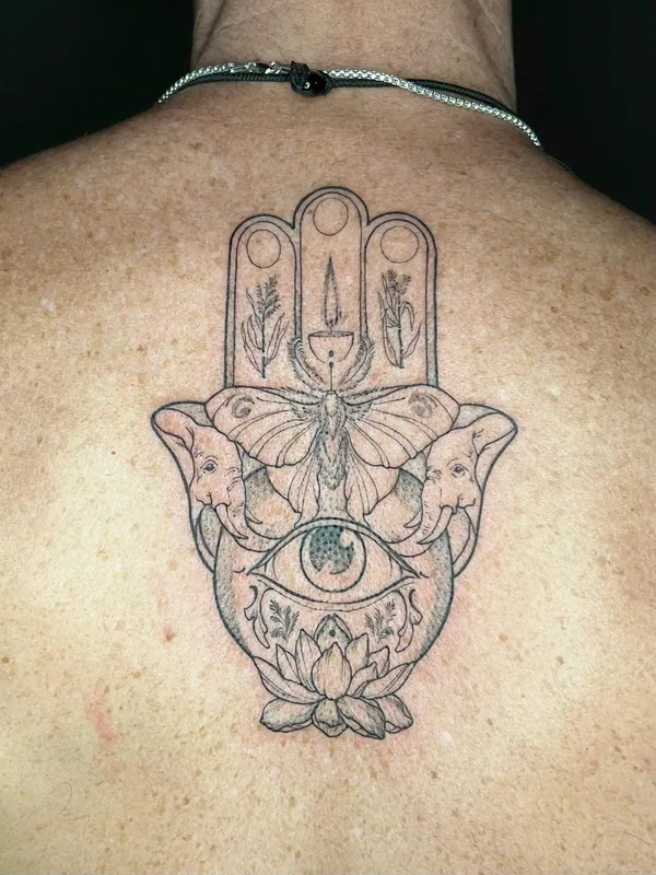 Fine-line hamsa tattoo containing elephants, a moth, a candle, an eye and a lotus centred on an upper back