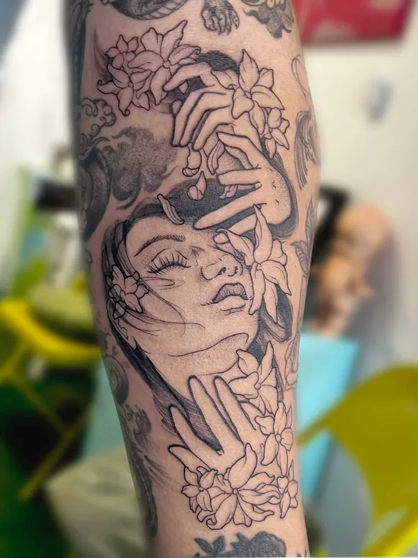 Black-and-grey tattoo of a woman's face with hands and jasmine blossoms drifting around it on a forearm