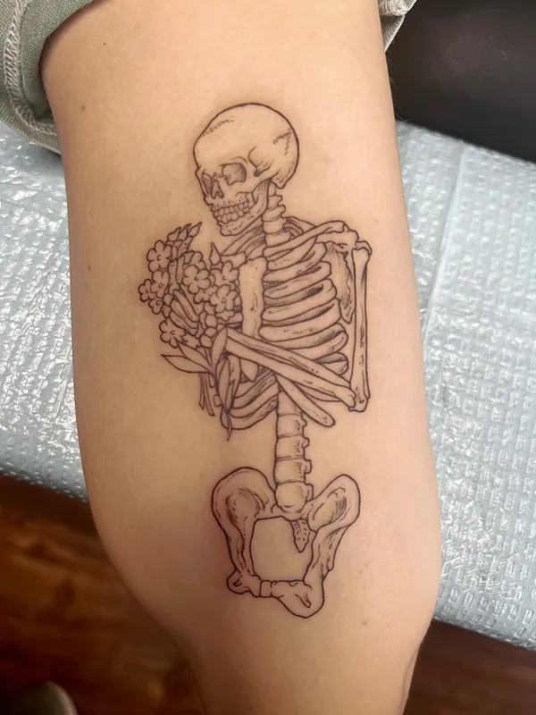 Fine-line skeleton tattoo from skull to pelvis holding a small bouquet of flowers against its ribs on a forearm