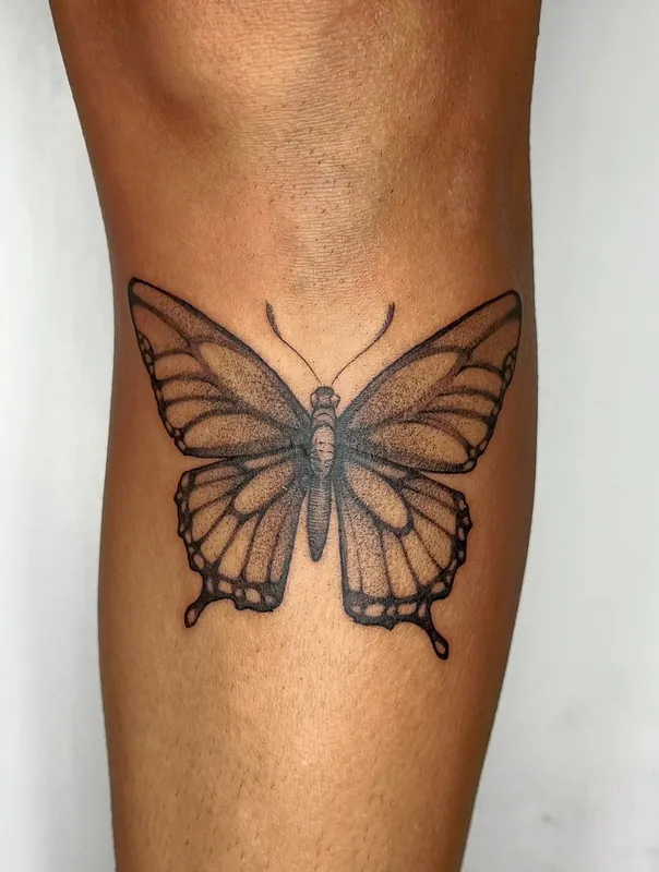 Black-and-grey dotwork swallowtail butterfly tattoo spread symmetrically below the back of a knee