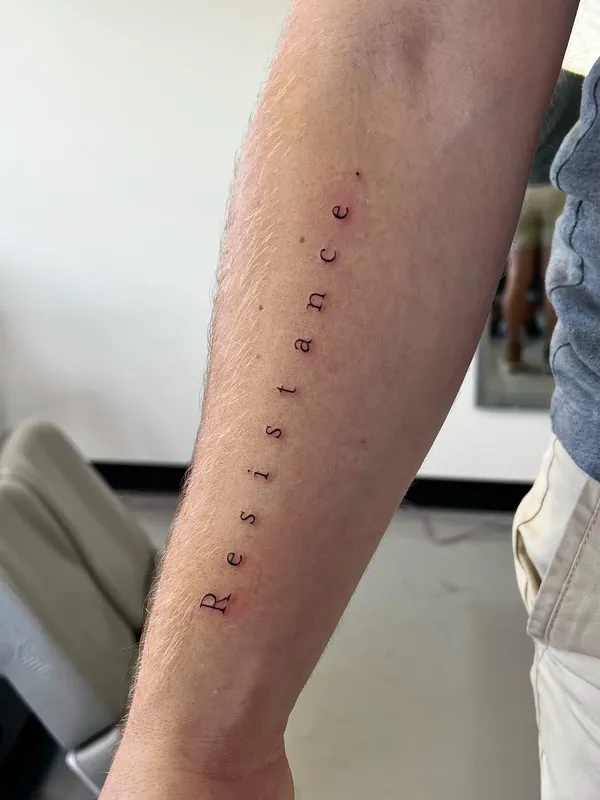 Fine-line serif lettering tattoo reading 'Resistance' running the length of an inner forearm