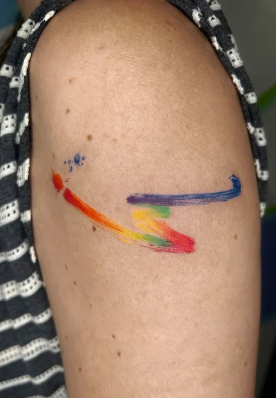 Color tattoo of two rainbow watercolor brushstrokes with a few paint splatters on an upper arm