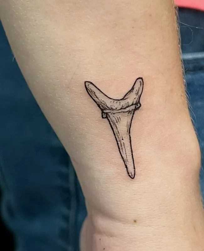 Small fine-line black-and-grey shark tooth tattoo with light hatching on the inside of a wrist