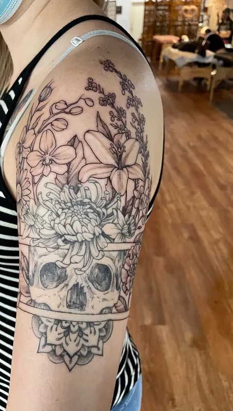 Black-and-grey tattoo of a skull beneath lilies, lilac and a chrysanthemum, split by a band with a mandala below, on an upper arm