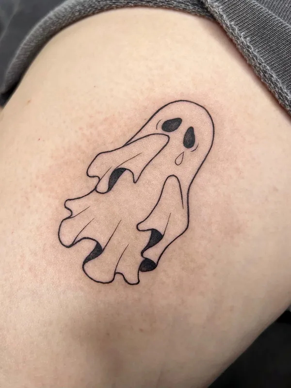 Bold linework tattoo of a small cartoon sheet ghost with hollow eyes and a single tear on a thigh