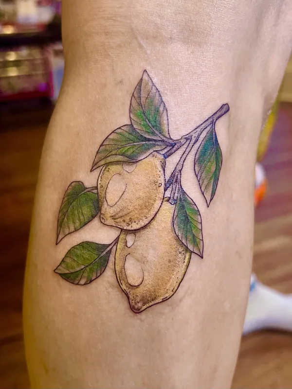 Color tattoo of two ripe lemons hanging from a leafy branch, shaded in yellow and green, on a calf
