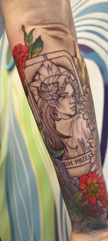Tarot-style High Priestess tattoo in black-and-grey framed by red and orange color zinnias on a forearm