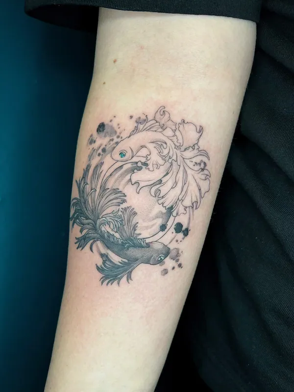 Fine-line tattoo of two betta fish circling in a yin-yang with ink splashes, one with a spot-color eye, on a forearm