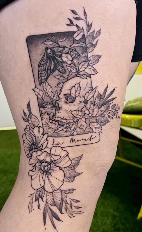 Black-and-grey tarot-style tattoo of a skull and moth in a card frame titled 'La Mort', with poppies, on a thigh