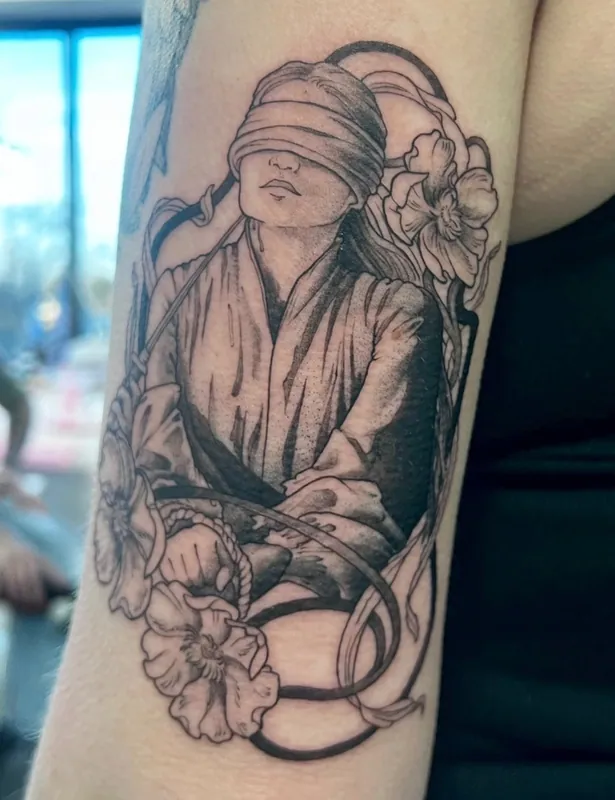 Black-and-grey tattoo of a blindfolded robed figure bound with rope inside an art nouveau frame of poppies on an inner arm