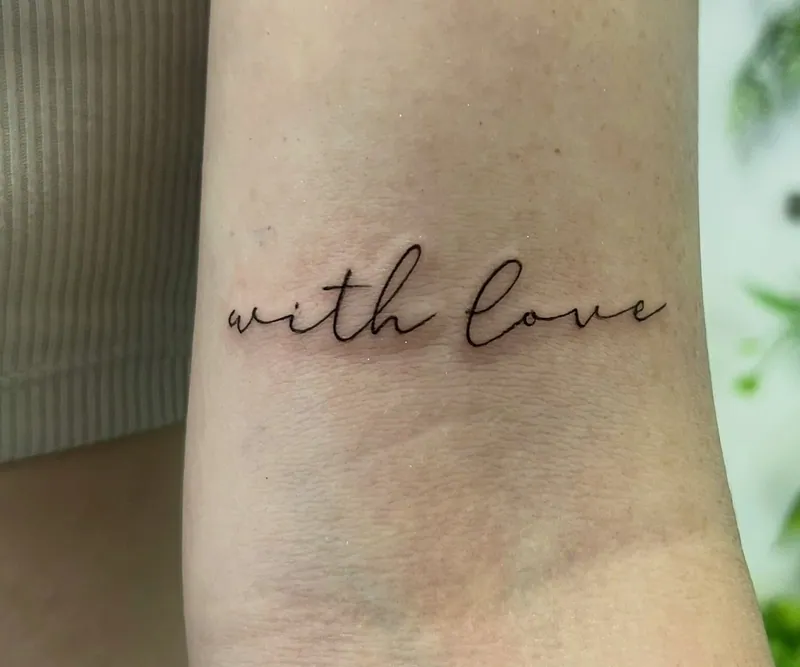 Fine-line cursive script tattoo reading 'with love' in a single flowing line on an inner upper arm