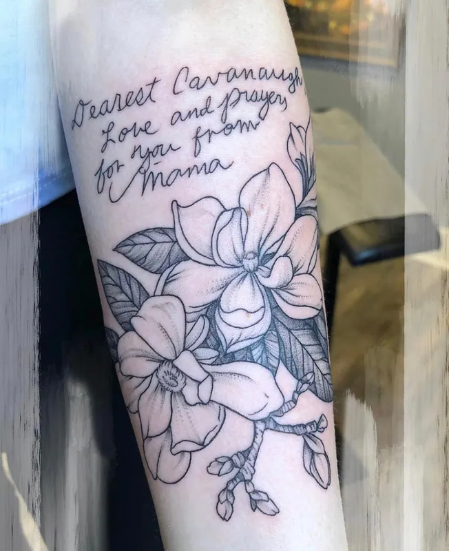 Black-and-grey magnolia tattoo beneath a handwritten note in a relative's script on a forearm