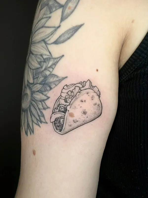 Small dotwork black-and-grey taco tattoo on the back of an upper arm beside an older sunflower piece