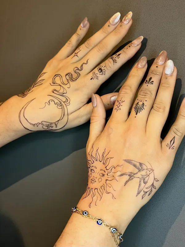 Fine-line hand tattoos on both hands: a crescent moon with a snake, a sun with a face, a swallow, an eye and finger ornaments