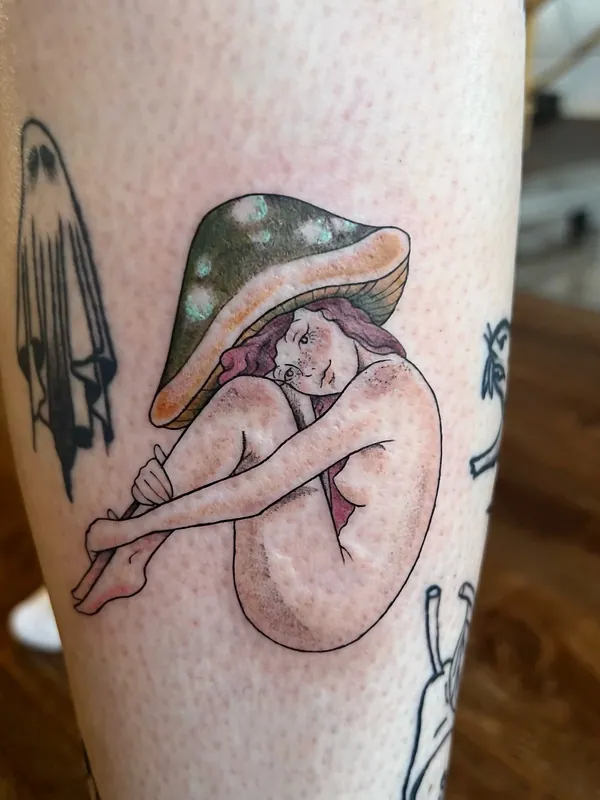 Color tattoo of a seated figure wearing a spotted mushroom cap, knees drawn up, on a calf
