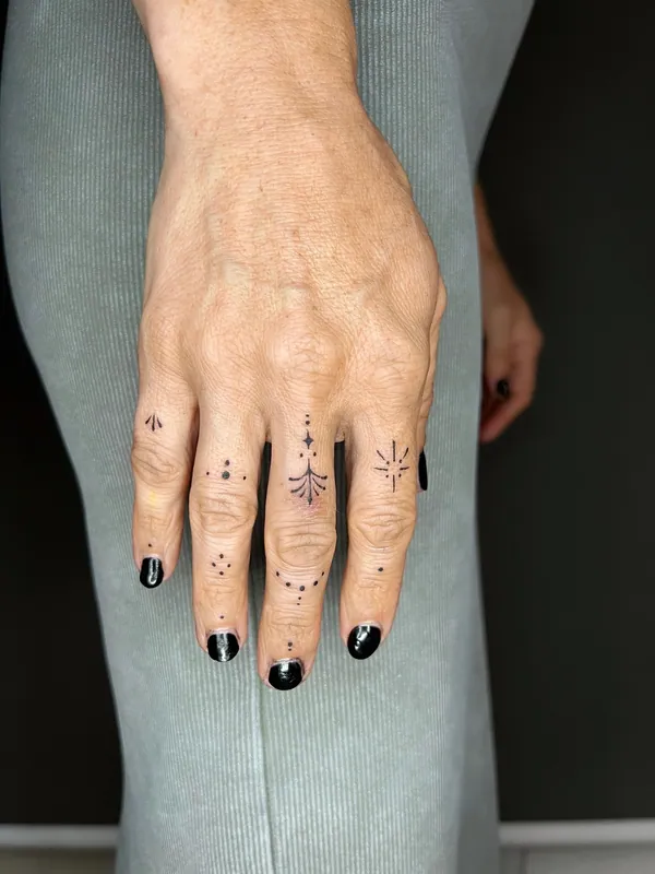 Tiny fine-line finger tattoos of dots, a sparkle, a small fern and a sunburst across four fingers
