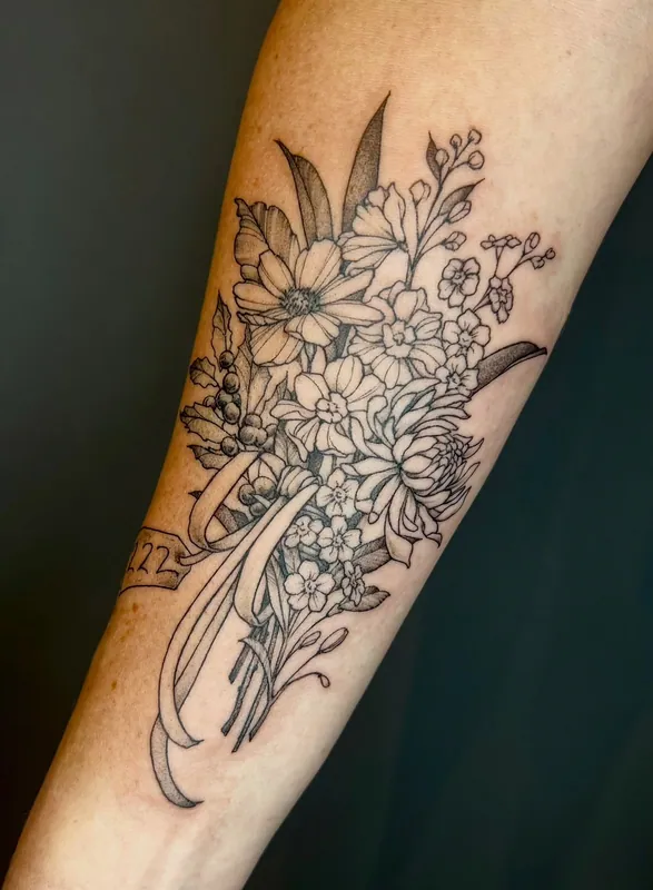 Fine-line black-and-grey bouquet tattoo of cosmos, chrysanthemum and wildflowers tied with ribbon and a small tag on a forearm