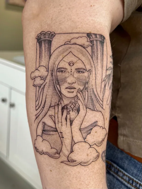Black-and-grey tattoo of a woman between stone columns holding a dark orb, with clouds and a crescent on her brow, on an inner arm