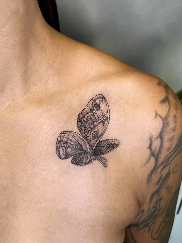 Black-and-grey realism tattoo of a butterfly with folded, patterned wings resting on a collarbone