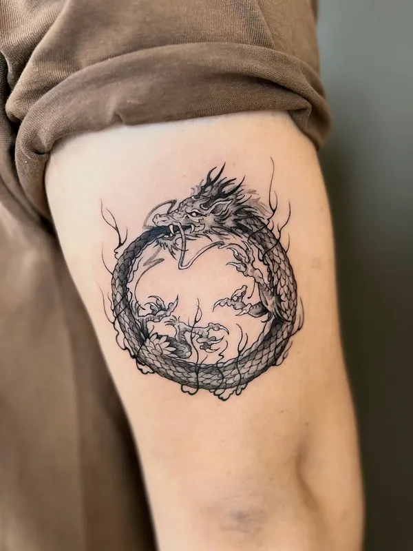 Fine-line black-and-grey ouroboros tattoo of an eastern dragon biting its own tail in a circle on an upper arm