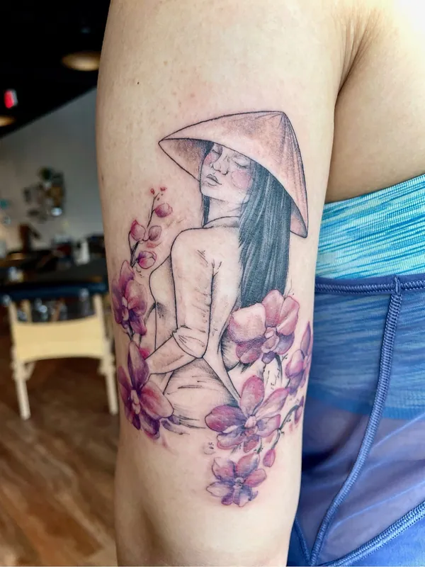 Tattoo of a woman in a conical hat seen from behind among pink color orchids on an upper arm